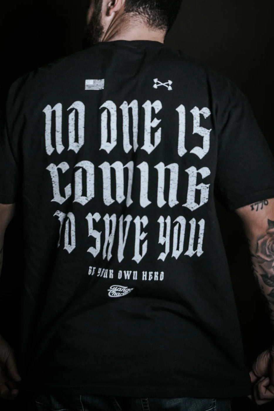A tattooed individual is seen from behind wearing Tango Charlie Apparel’s "No One Is Coming To Save You - Premium Tee" with bold white motivational text, standing out against a dark background.