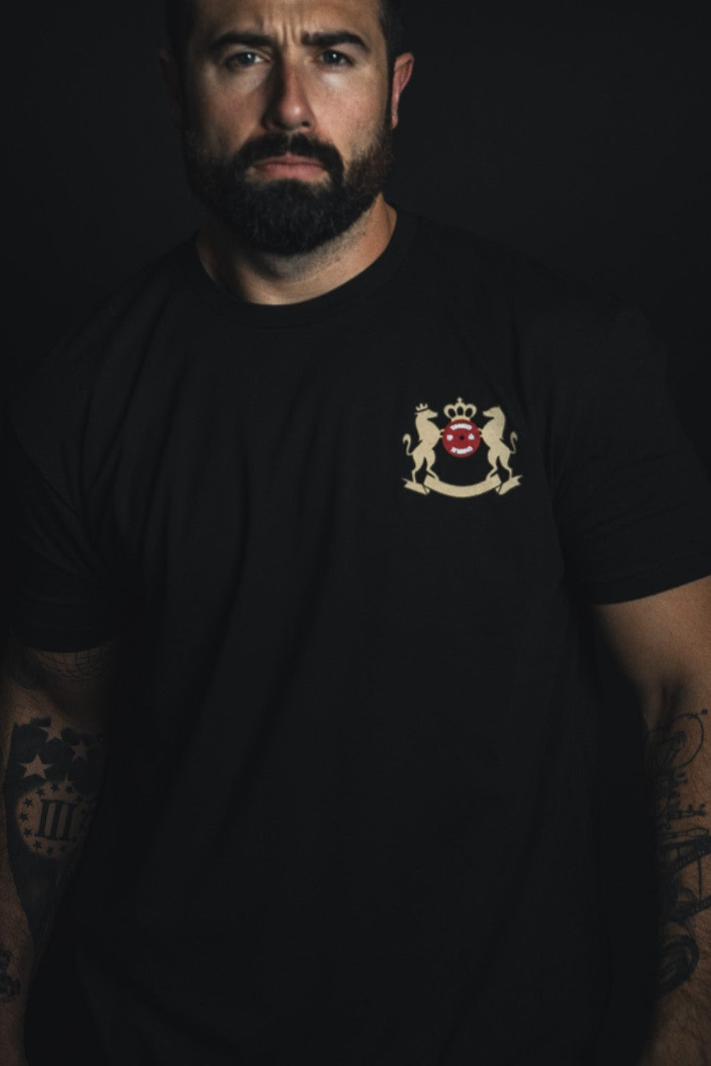 A bearded man stands against a dark backdrop, wearing the Deadlift Addiction Tee by Tango Charlie Apparel, featuring a crest with a red lion and mythical creatures. Tattoos snake down his arms as he gives you a serious look, echoing the lifter generals warning to challengers.