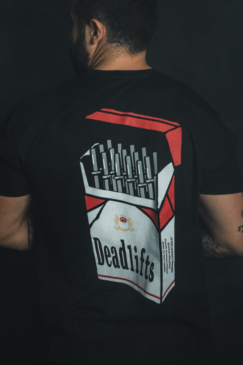 A man showcases his love for lifting in a Deadlift Addiction - Tee by Tango Charlie Apparel. The tee, with its Deadlifts cigarette pack design of mini barbells in red, white, and black, strikingly contrasts his tattooed arms against a dark background—a true lifter generals warning.