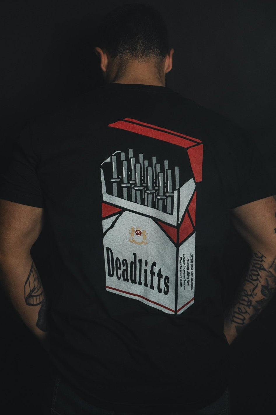 A tattooed individual turns away from the camera, wearing Tango Charlie Apparels Deadlift Addiction - Tee, a black shirt featuring a cigarette pack graphic labeled Deadlifts with a humorous lifter generals warning.
