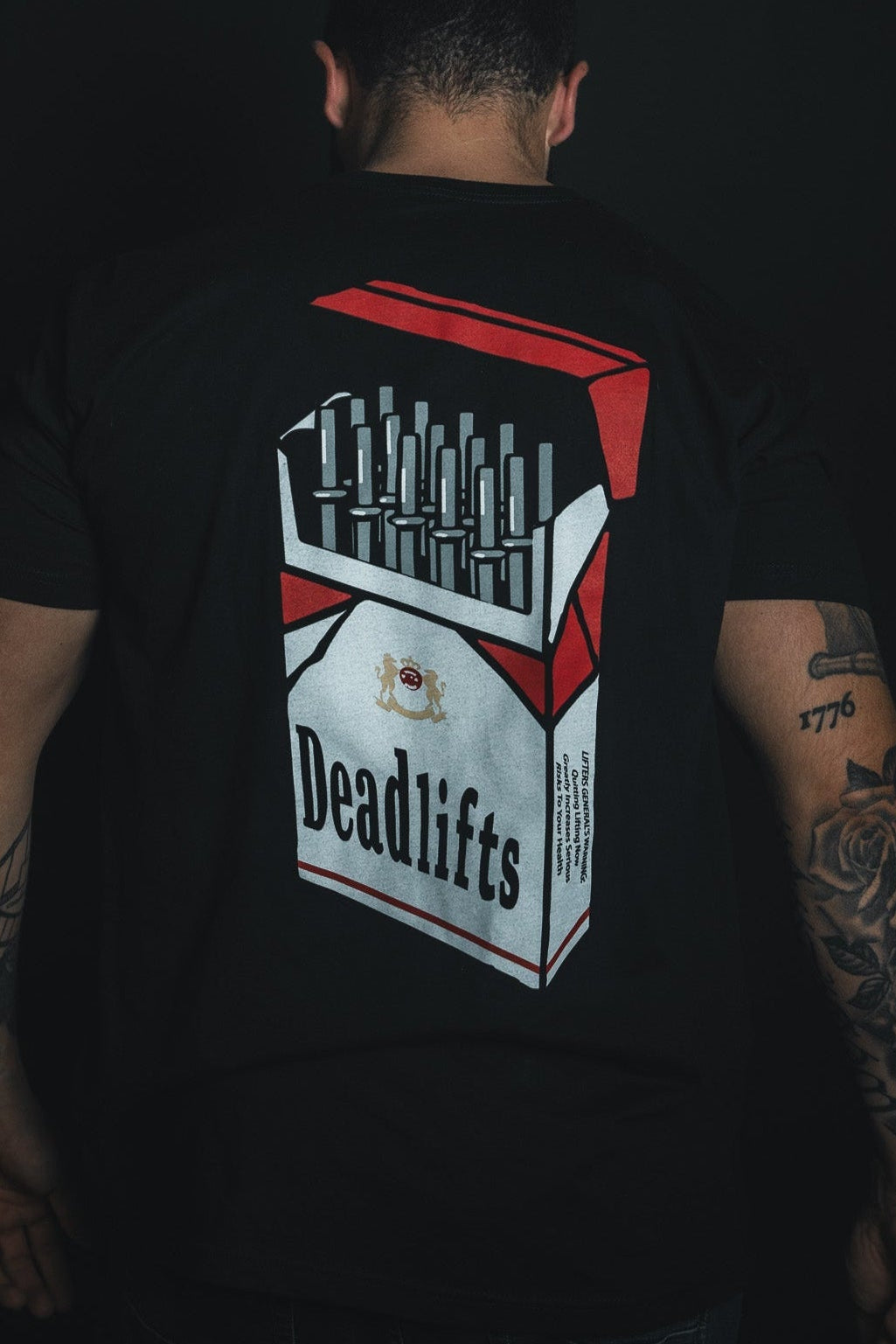A person faces away wearing the Deadlift Addiction Tee from Tango Charlie Apparel, showcasing a cigarette pack graphic labeled Deadlifts, filled with barbells. Tattoos on their arms warn of the deadlift lifestyles power and allure.