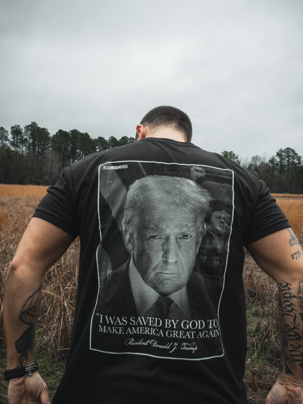 A man stands outdoors, photographed from behind, wearing the Born United Private Label "Legend - Saved" black T-shirt featuring Donald J. Trump’s face and the phrase: “I was saved by God to Make America Great Again.”.