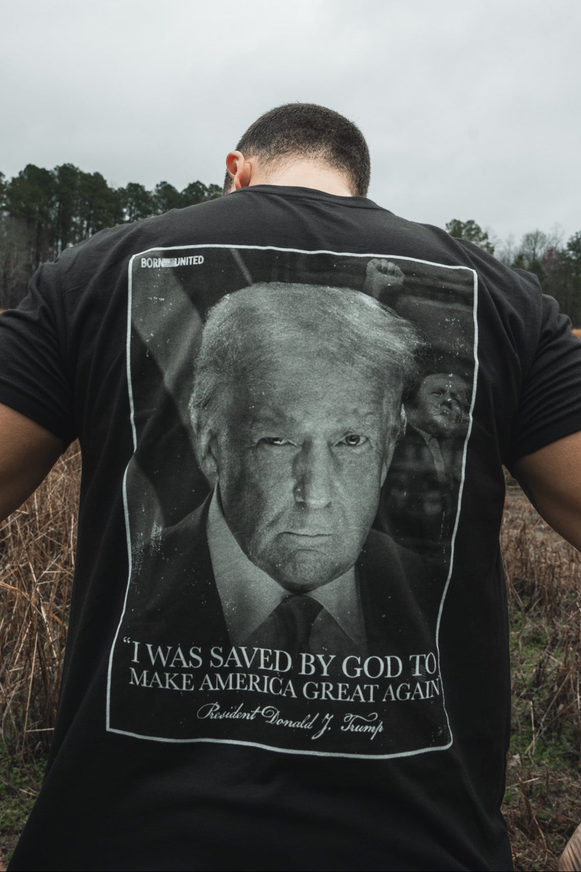 A man stands outdoors, photographed from behind, wearing the Born United Private Label "Legend - Saved" black T-shirt featuring Donald J. Trump’s face and the phrase: “I was saved by God to Make America Great Again.”.