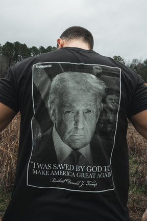 A man stands outdoors, photographed from behind, wearing the Born United Private Label "Legend - Saved" black T-shirt featuring Donald J. Trump’s face and the phrase: “I was saved by God to Make America Great Again.”.