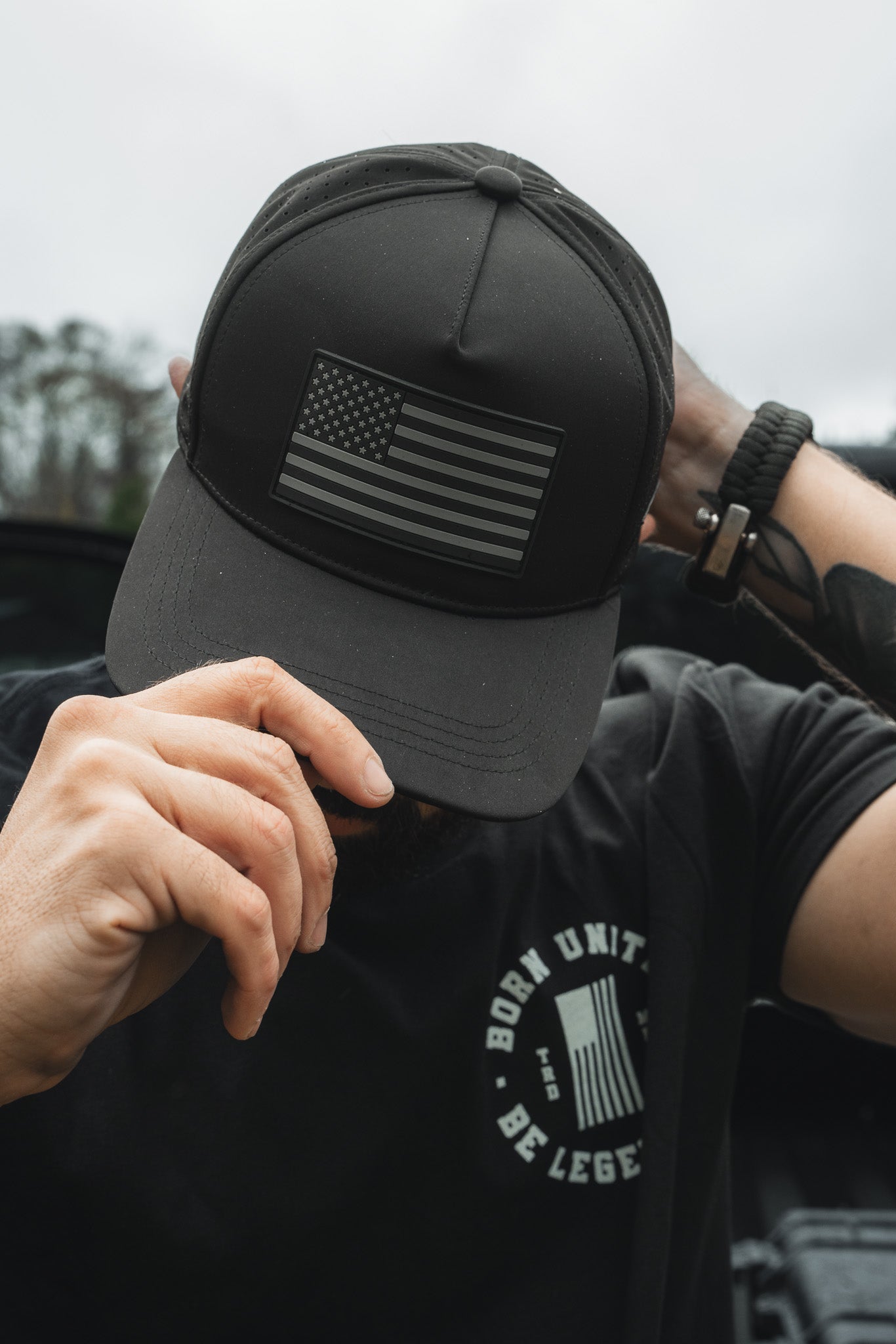 Person adjusts a Born United Premium Flag Snapback - Blackout, displaying a monochrome American flag patch. They sport a black T-shirt, watch, and visible tattoos on their arm. The blurred backdrop features trees.