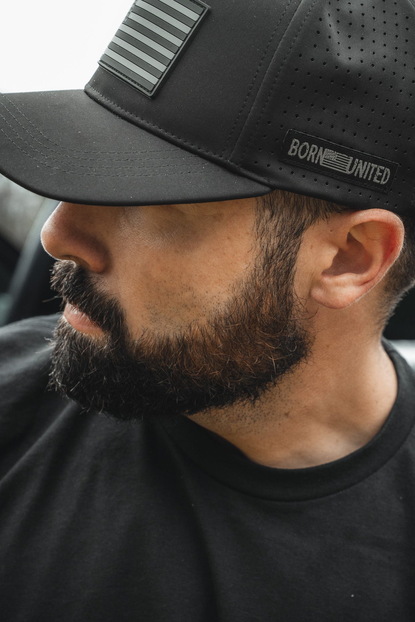 A bearded man wears a Born United Premium Flag Snapback - Blackout by Born United Private Label and a T-shirt, featuring perforations, BORN UNITED, and a stylized American flag near the brim, with his face partially turned from the camera.