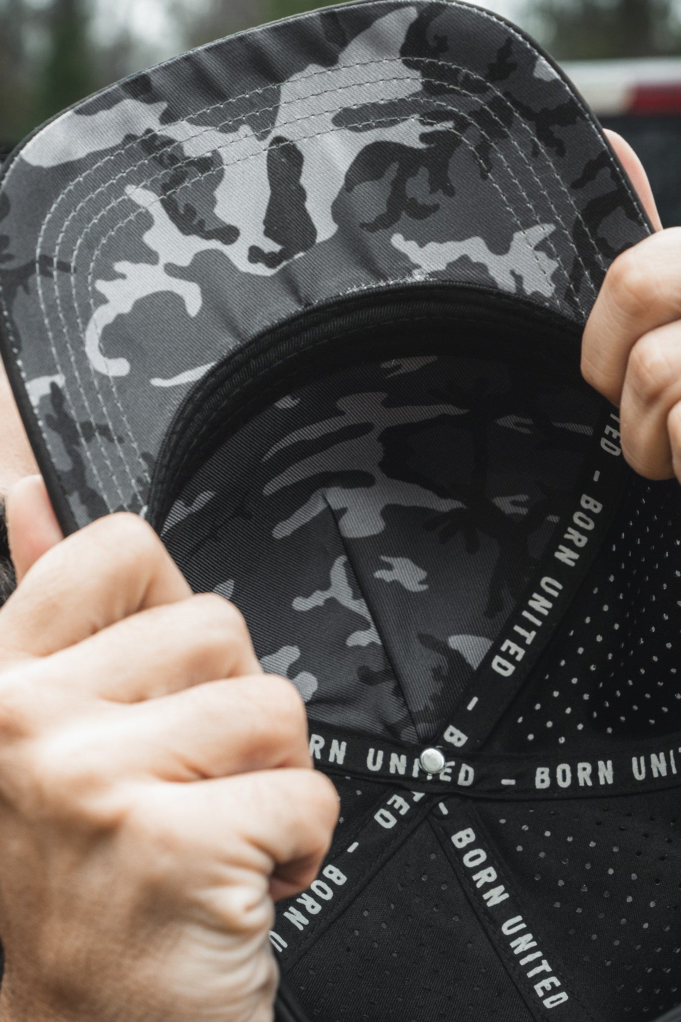 A person grips the brim of a camo-patterned Born United Premium Flag Snapback - Blackout, subtly concealing their face. The inner band reads BORN UNITED, while the blurred background accentuates the caps intricate details.