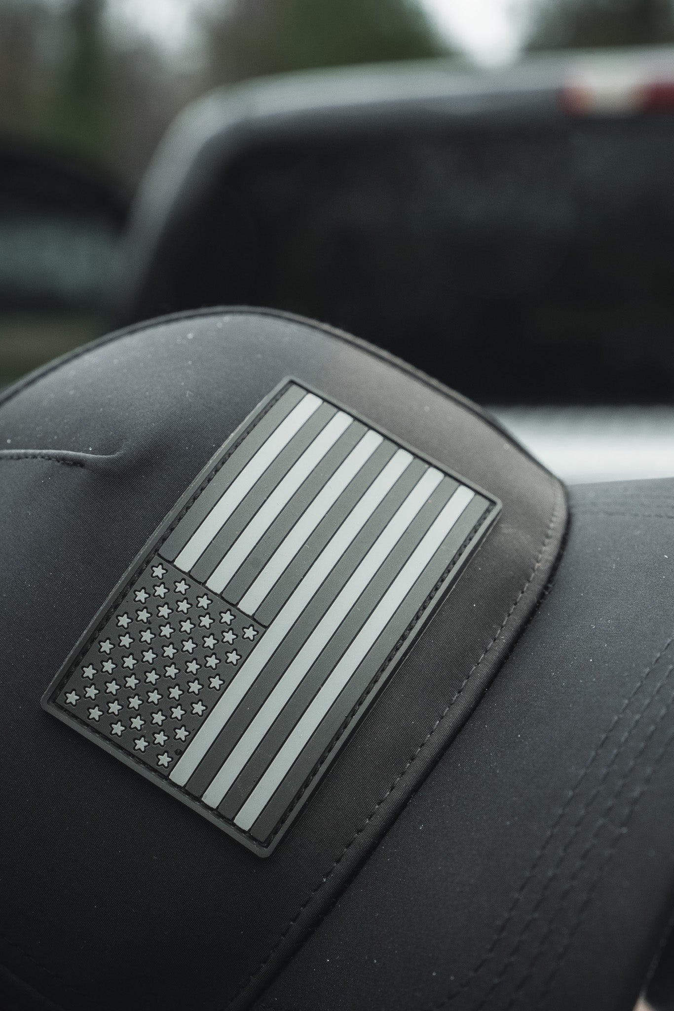 Close-up of the Born United Premium Flag Snapback - Blackout by Born United Private Label, showcasing a black-and-white American flag patch on grey. The caps brim is in focus with a blurred vehicle in the background.