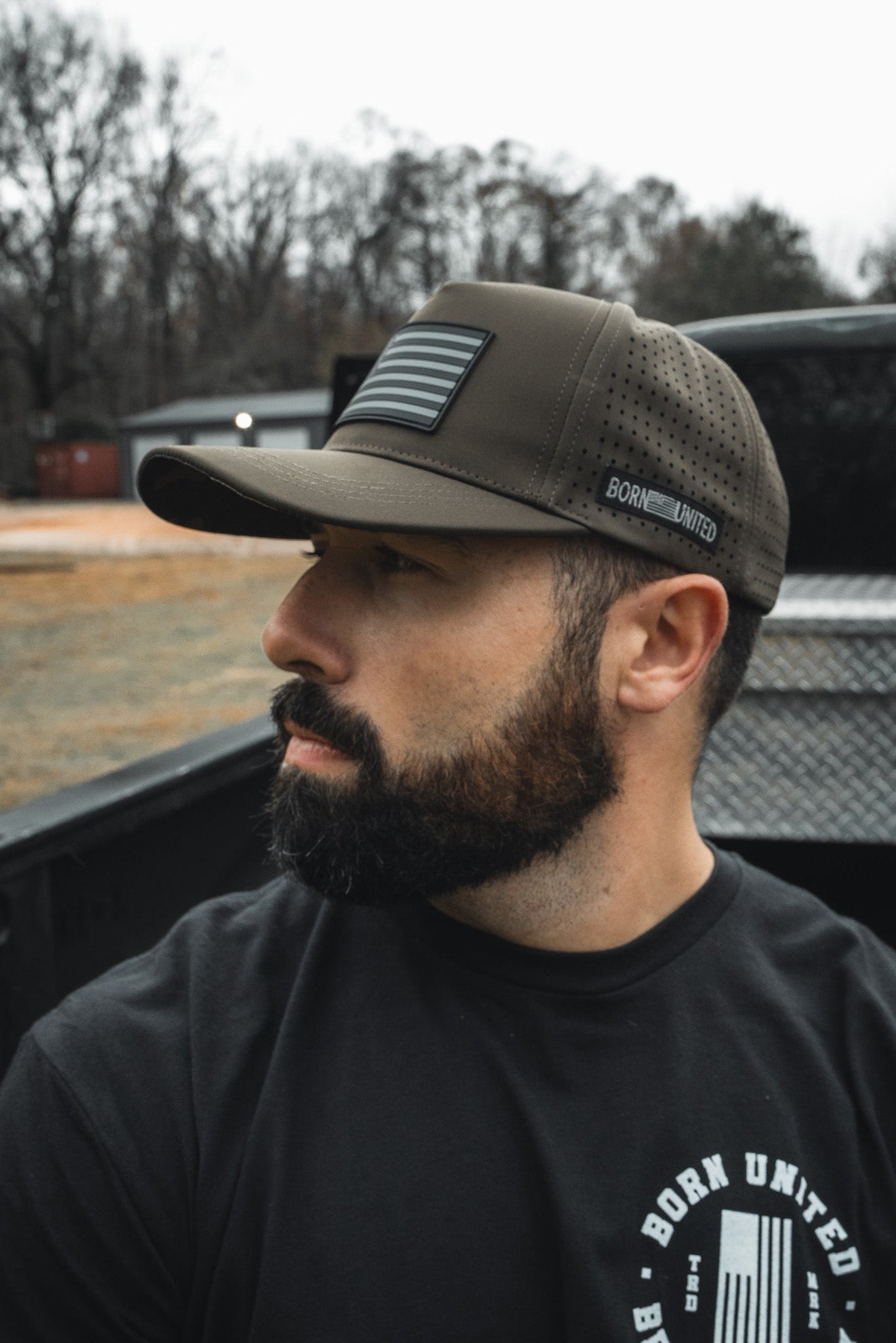 A bearded man in a black shirt with a circular logo sits in the back of a pickup truck, wearing the Born United Premium Flag Snapback - Olive by Born United Private Label. The camo print cap suits the rugged landscape where trees meet a cloudy sky.