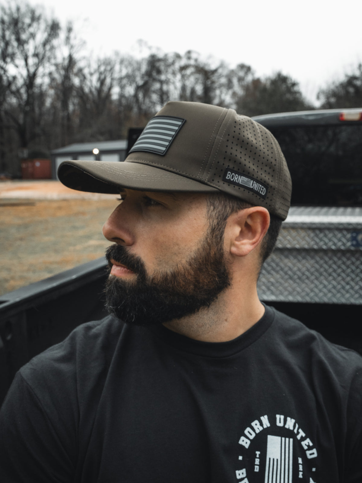 A bearded man in a black T-shirt and an olive green "Legend - Saved" baseball cap by Born United Private Label with an American flag patch sits in a pickup truck bed, looking to the side. Trees and a building blur in the background.