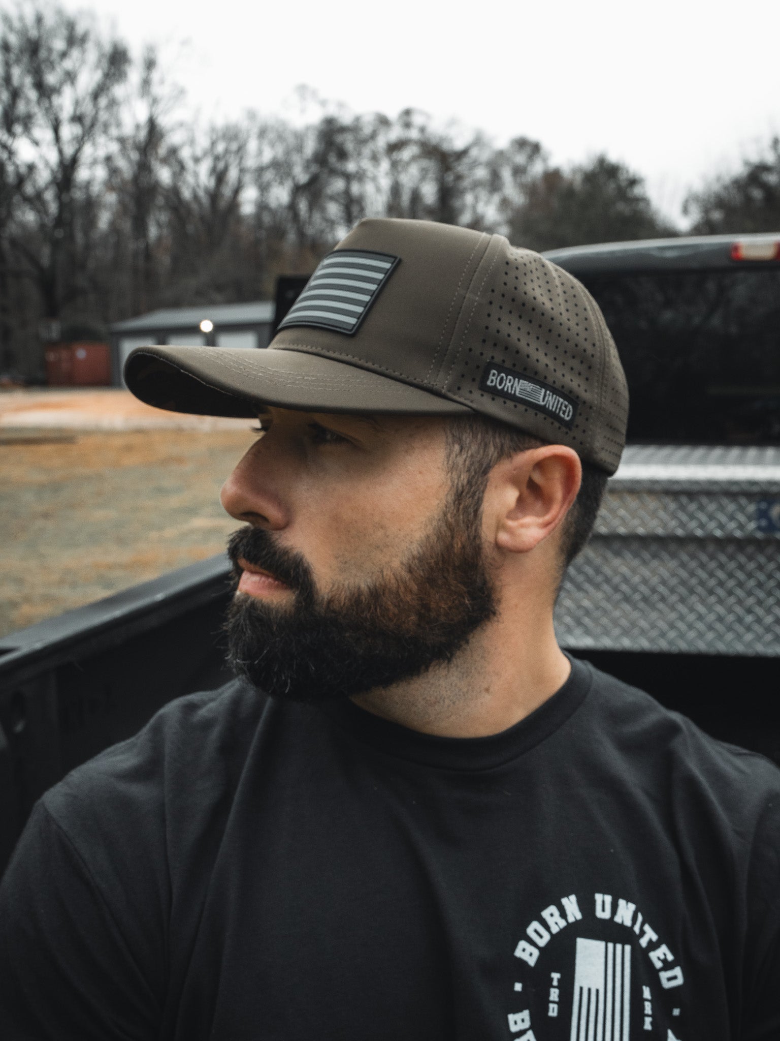 A bearded man in a black T-shirt and an olive green "Legend - Saved" baseball cap by Born United Private Label with an American flag patch sits in a pickup truck bed, looking to the side. Trees and a building blur in the background.