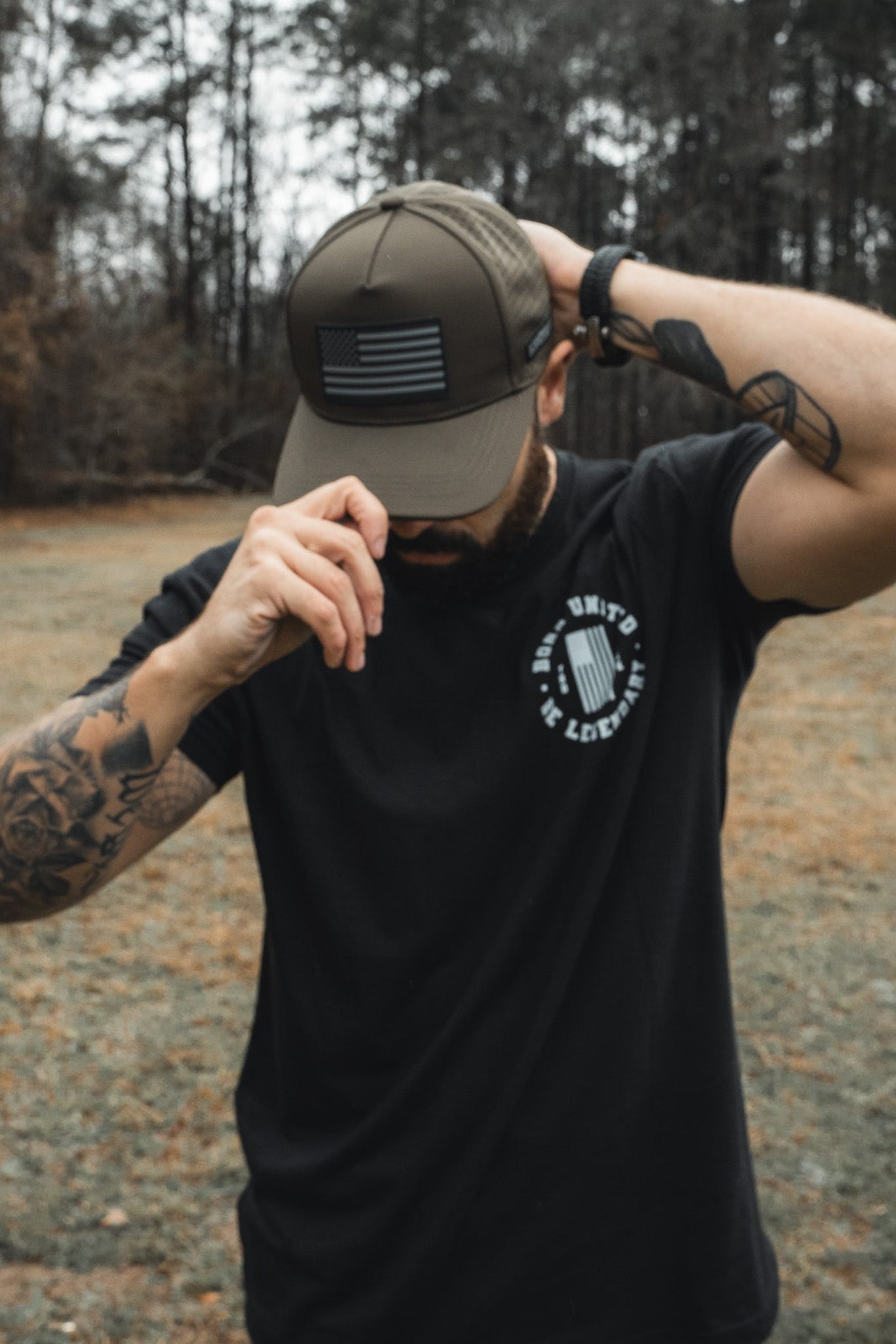 A man in a black t-shirt and camo cap stands in a grassy field, trees behind him. Tattoos cover his right arm as he adjusts the Born United Premium Flag Snapback - Olive featuring a black and white American flag design, with his left hand.