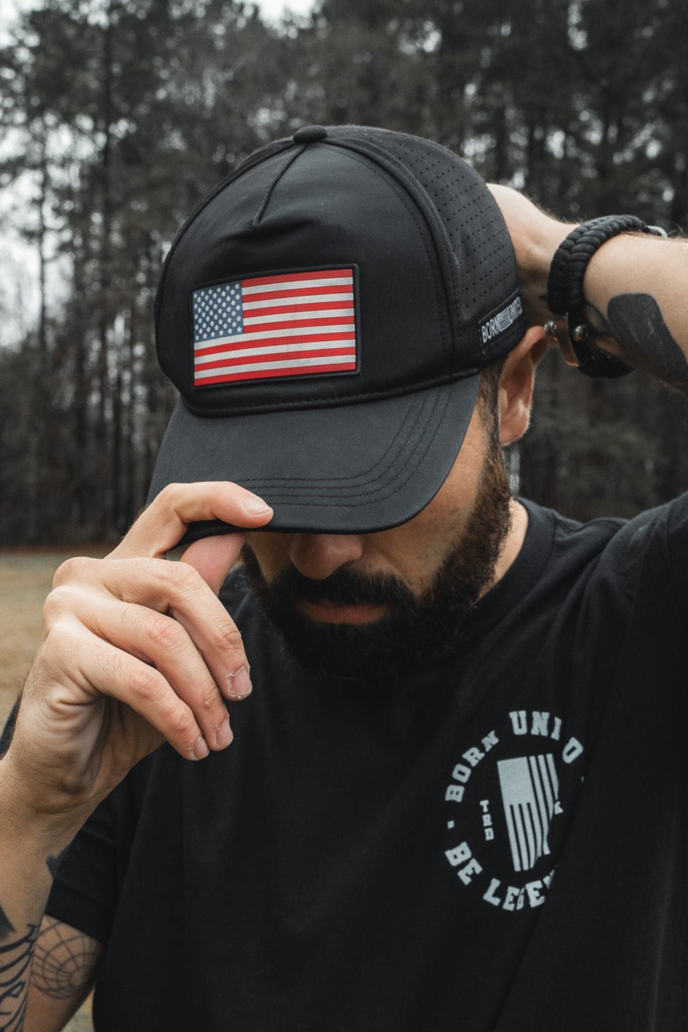 A bearded man, wearing the Born United Premium Flag Snapback - RW&B from Born United Private Label adjusts it outdoors. He sports a black T-shirt with white text and has tattoos on his arm, set against trees and a grassy field backdrop.