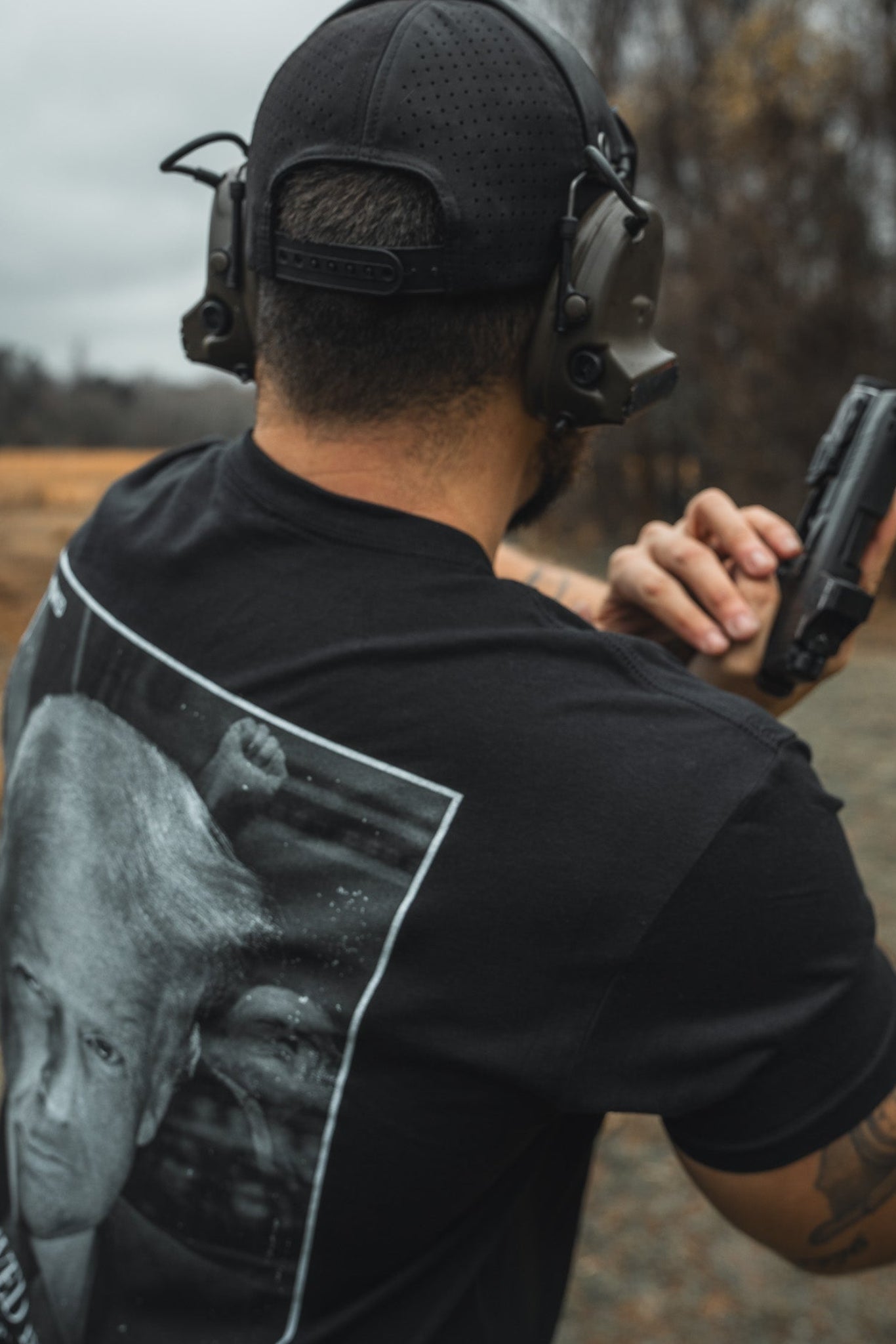 A person wearing a Born United Premium Flag Snapback - RW&B by Born United Private Label and a headset is at an outdoor shooting range, holding a handgun. Their shirt features a printed image against trees and an overcast sky, creating a distinctly American atmosphere.