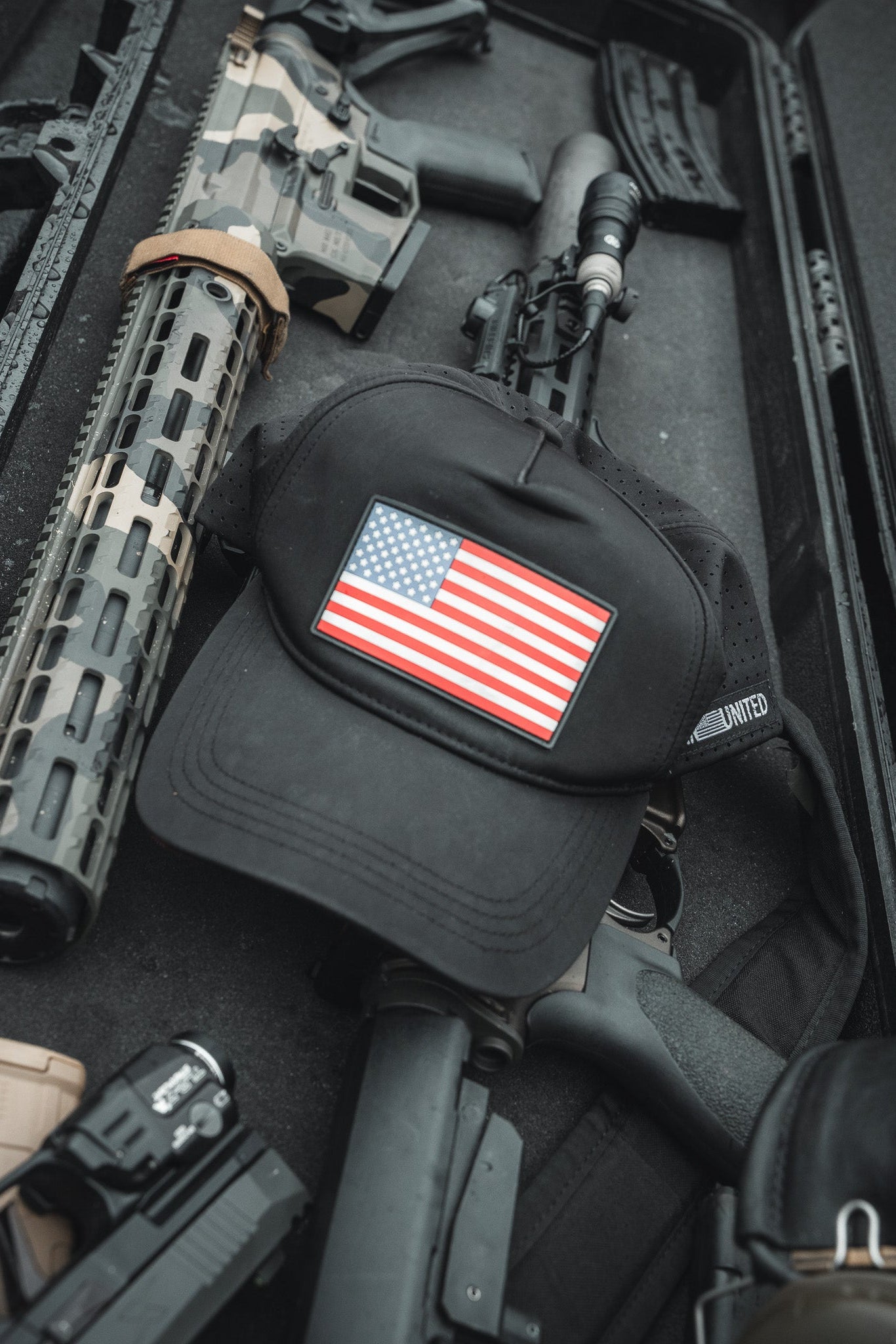A Born United Premium Flag Snapback - RW&B sits atop a sleek black case filled with firearms and accessories, including rifles and a handgun with camouflage accents. This display from Born United Private Label embodies rugged patriotism.
