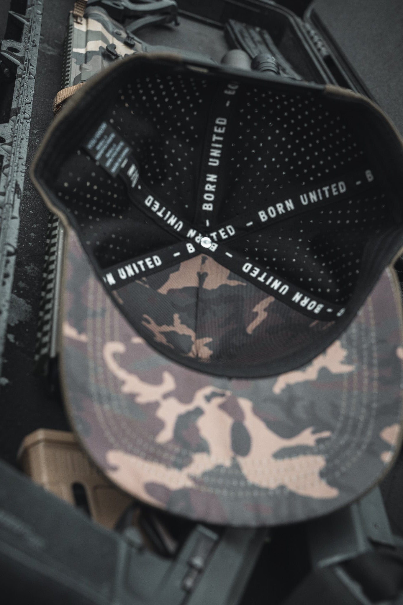 The Born United Premium Flag Snapback - Olive, a handmade camo cap featuring BORN UNITED text inside, rests on a rifle case alongside tactical accessories, embodying a military-inspired theme.