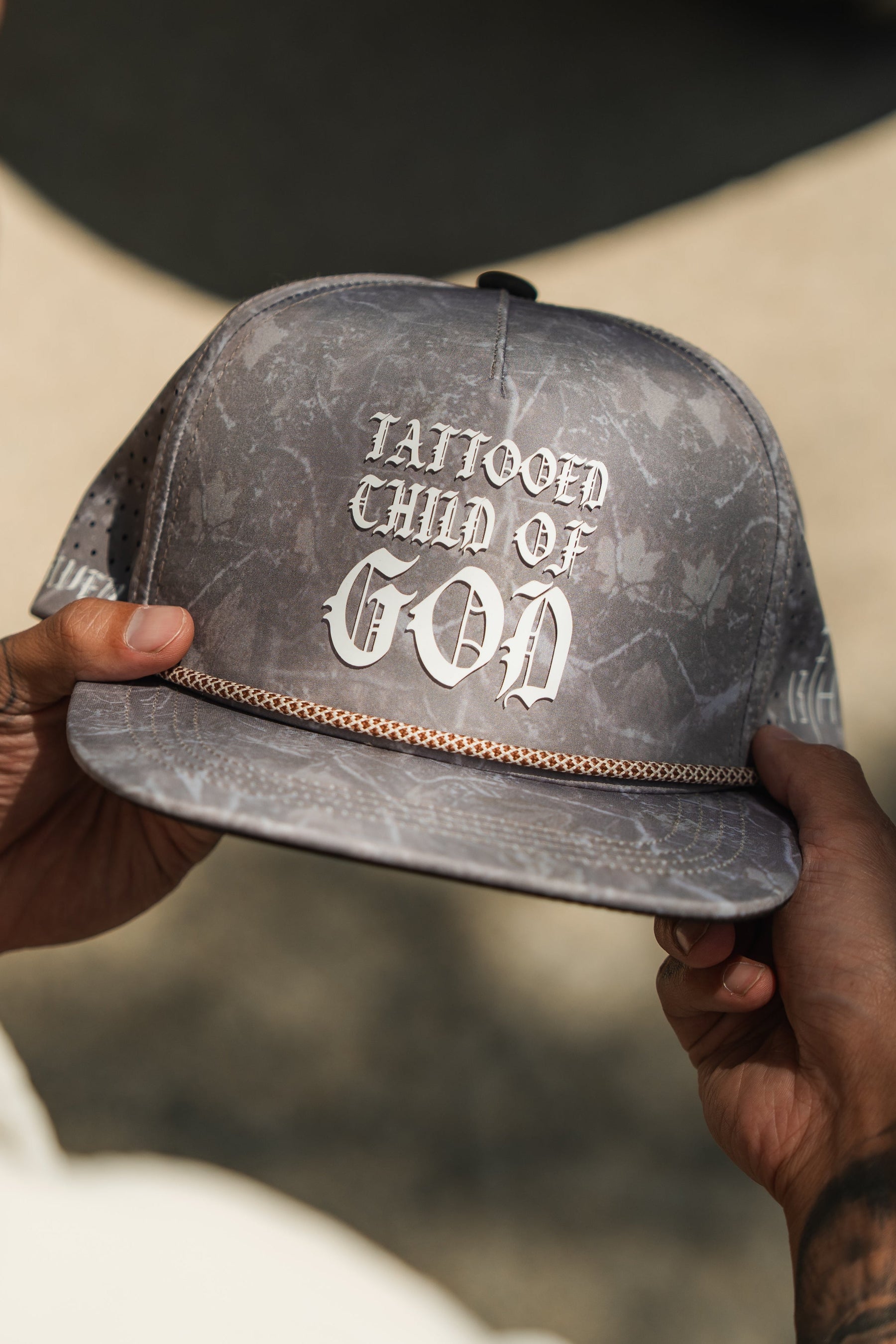 A person holds a Relentless Betrayal Tattooed Child of God Deadwood Camo PVC Snapback, featuring bold white gothic lettering and a chain detail above the brim.