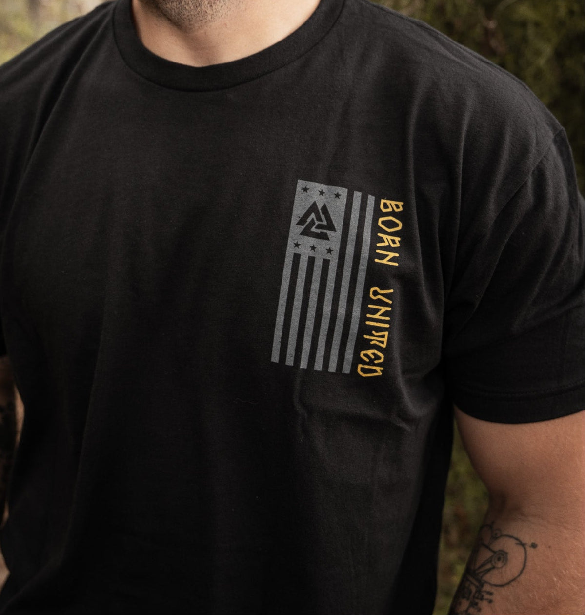 In a wooded area, a bearded man wearing a black Warrior Spirit T-shirt by Born United Private Label stands, evoking the strength of First Responders. He looks to the side, surrounded by trees and fallen leaves that symbolize resilience.
