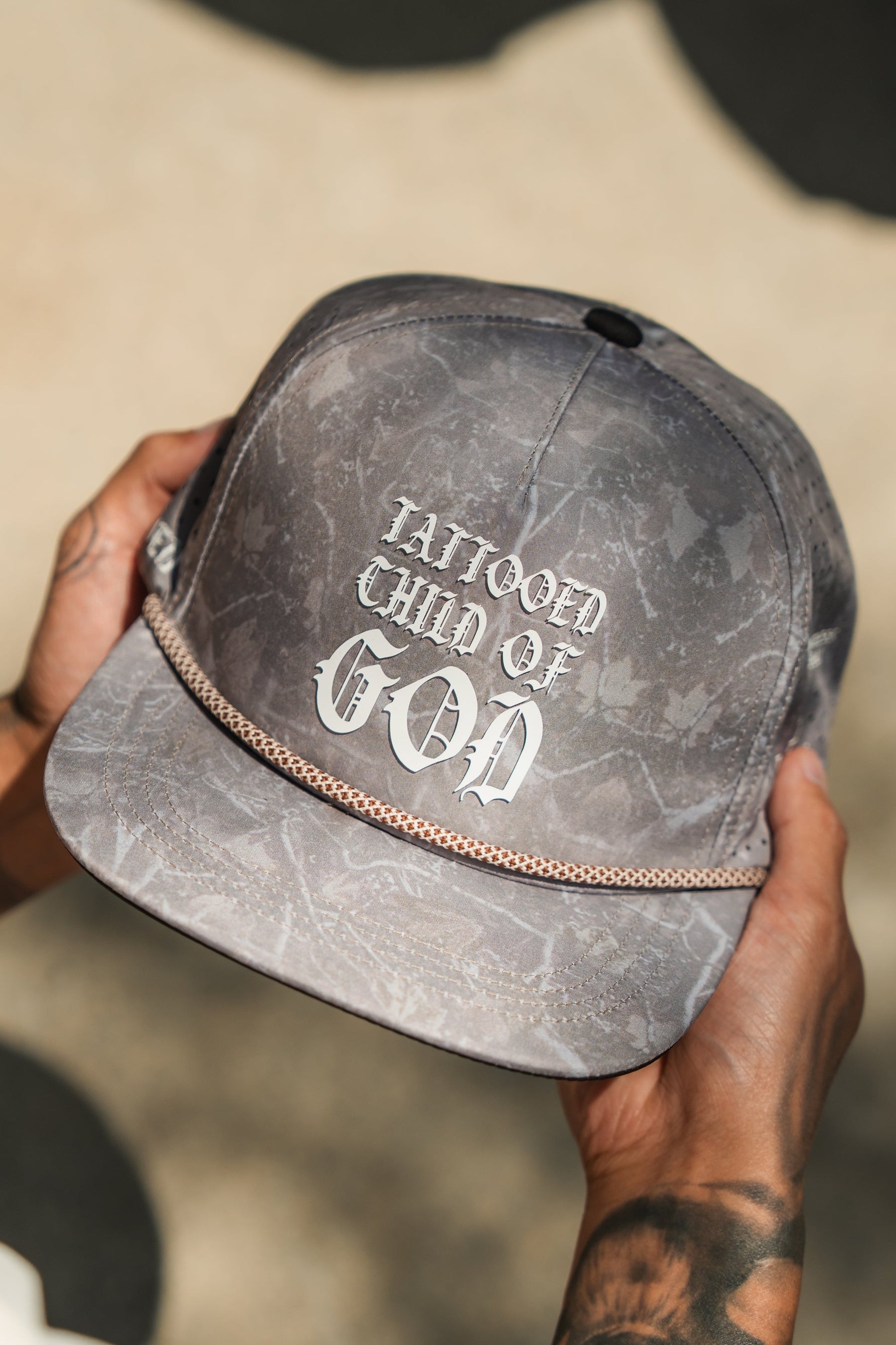 A person with tattooed hands holds a Tattooed Child of God Deadwood Camo PVC Snapback by Relentless Betrayal—a gray cap with white gothic text and a decorative chain across the brim.