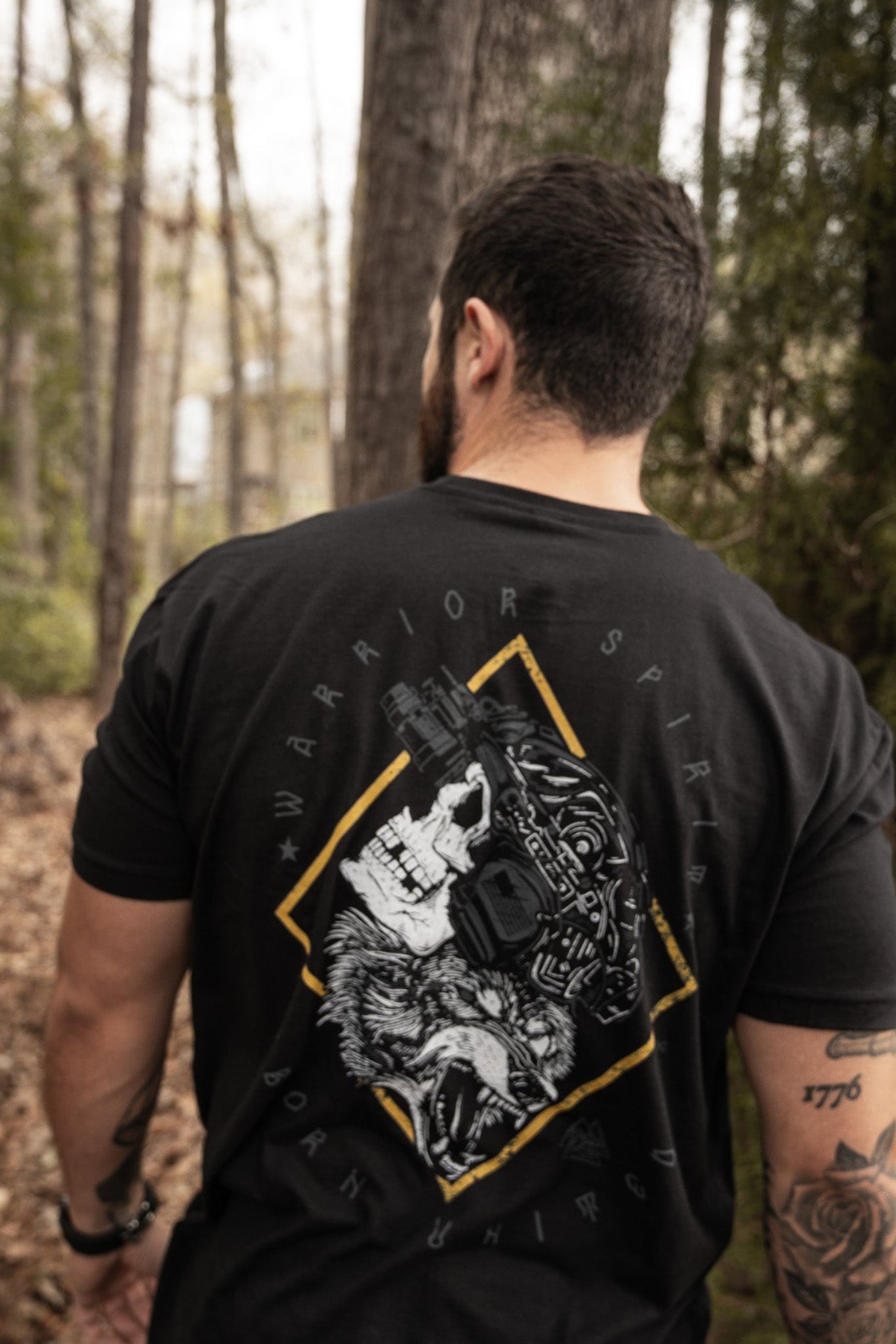 A tattooed person walks in a forest, wearing a Born United Private Labels Warrior Spirit black T-shirt. The back features a skull and wolf graphic, symbolizing the strength of first responders with the words Warrior Spirit.