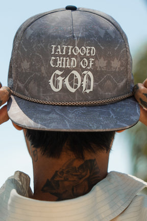 Wearing a light shirt and showing neck tattoos, a person stands outdoors under blue skies holding the Relentless Betrayal Tattooed Child of God Deadwood Camo PVC Snapback, showcasing bold Christian style.