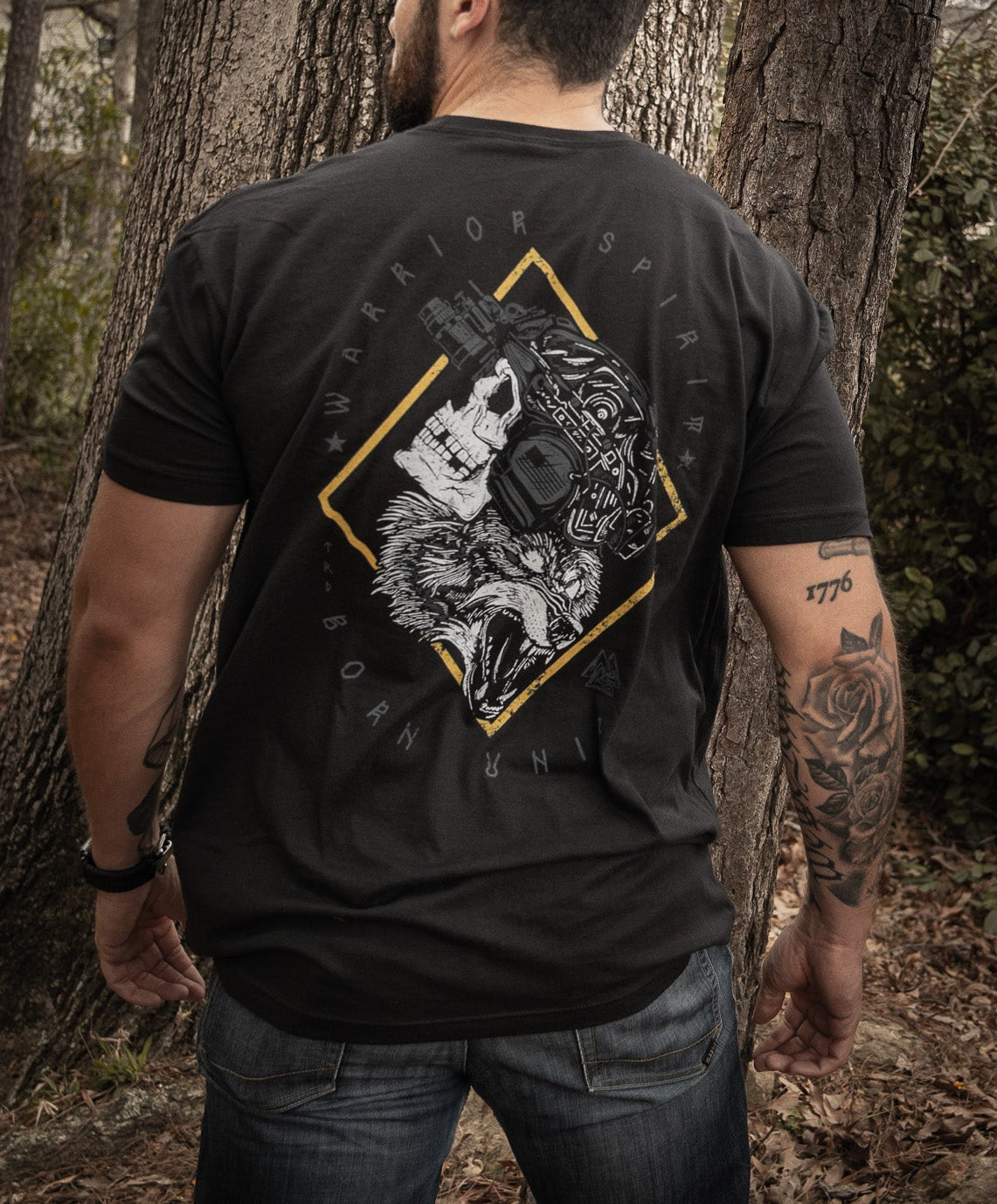 A man stands in a forest with his back to the camera, wearing Born United Private Labels Warrior Spirit black t-shirt, featuring a wolf graphic and text. His tattooed left arm complements the strength of character amidst tall trees and fallen leaves.