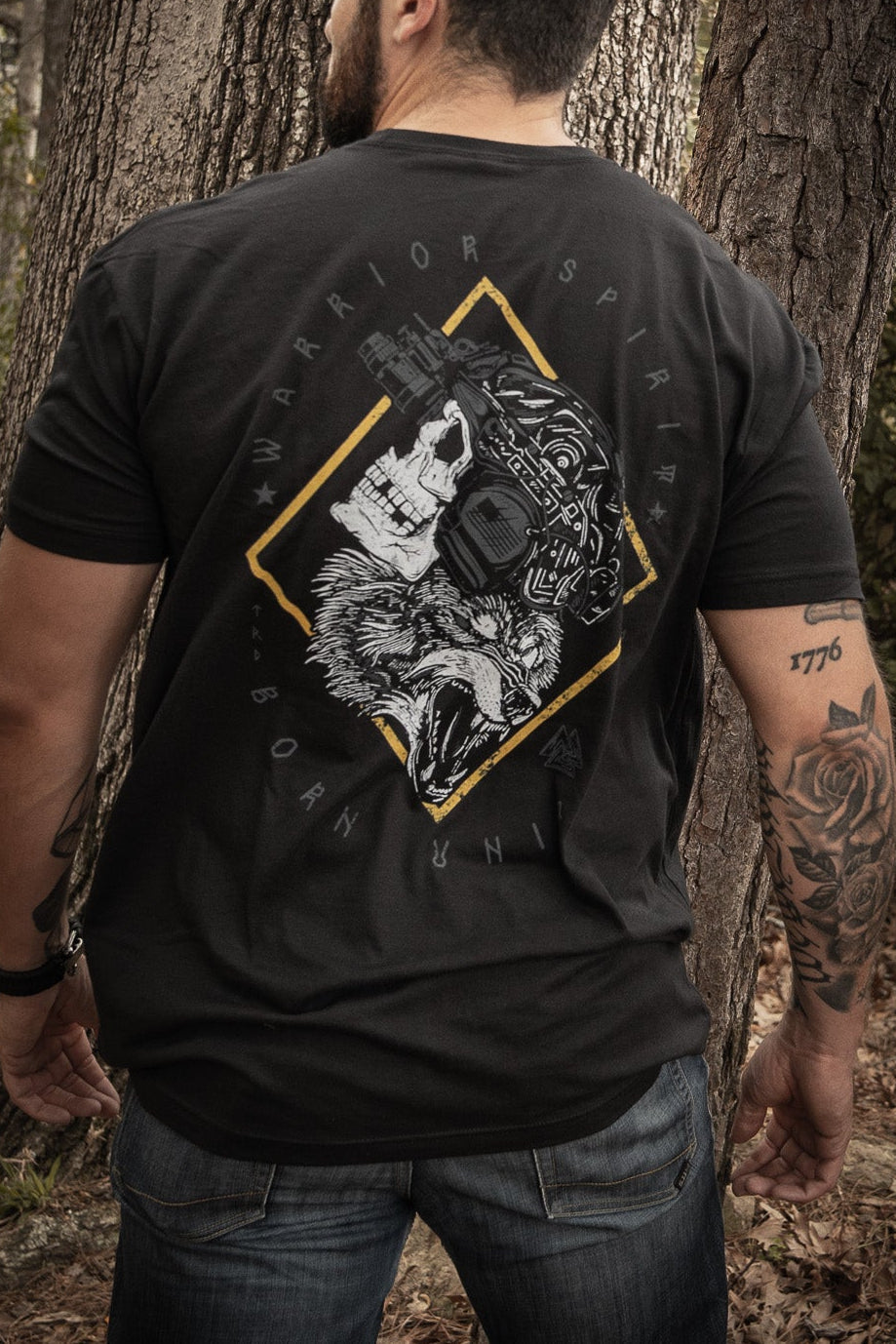 A man stands in a forest with his back to the camera, wearing Born United Private Labels Warrior Spirit black t-shirt, featuring a wolf graphic and text. His tattooed left arm complements the strength of character amidst tall trees and fallen leaves.
