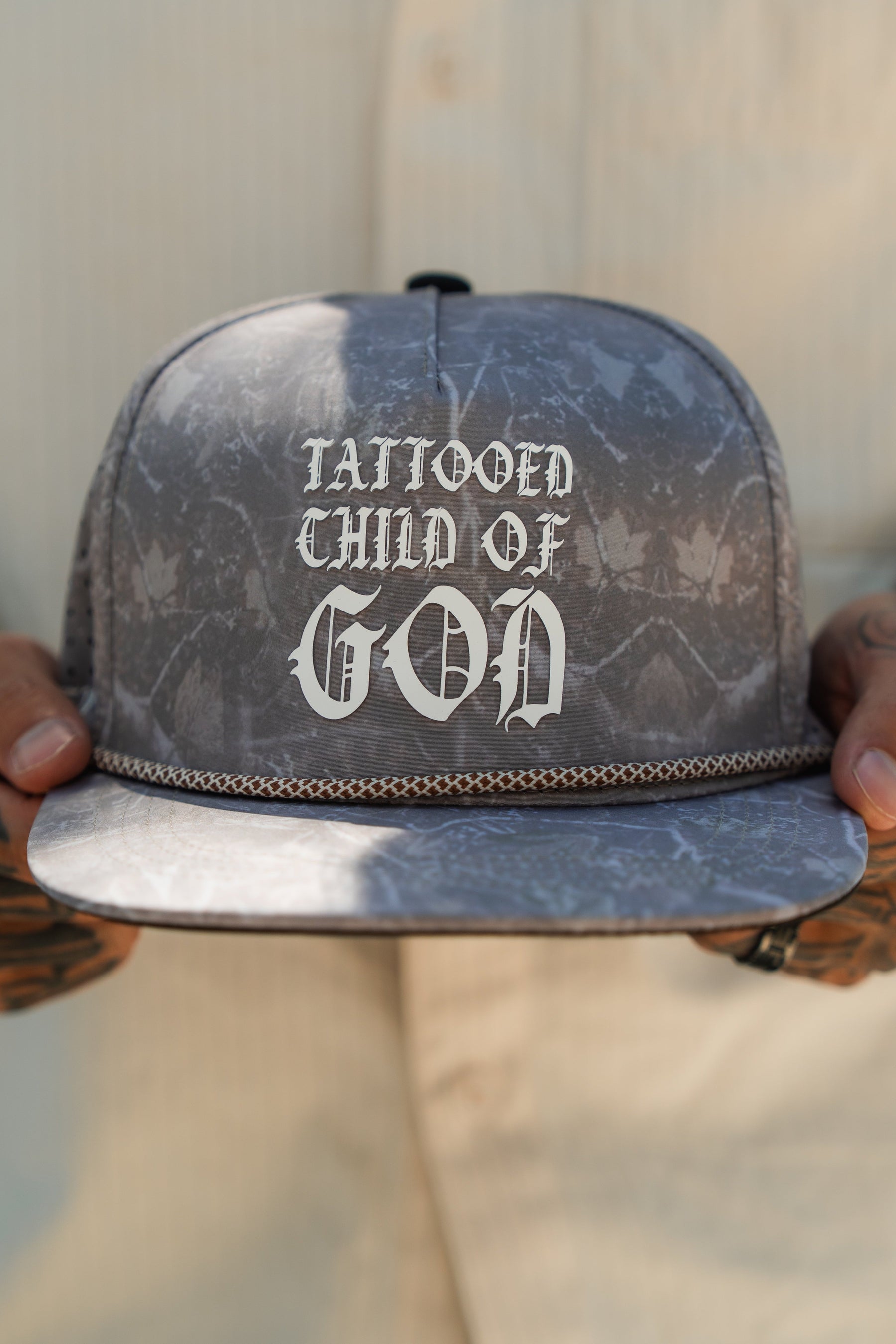 A person with tattooed hands holds a Relentless Betrayal Tattooed Child of God Deadwood Camo PVC Snapback, featuring bold white gothic text. The light shirt and subtle background highlight this unique faith-inspired hat.