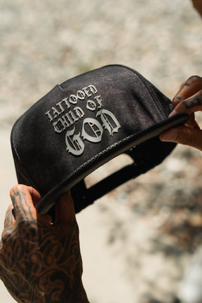 A person with tattooed hands holds a Tattooed Child of God Mineral Wash PVC Snapback by Relentless Betrayal, featuring bold white gothic letters on the front, against a blurred background.
