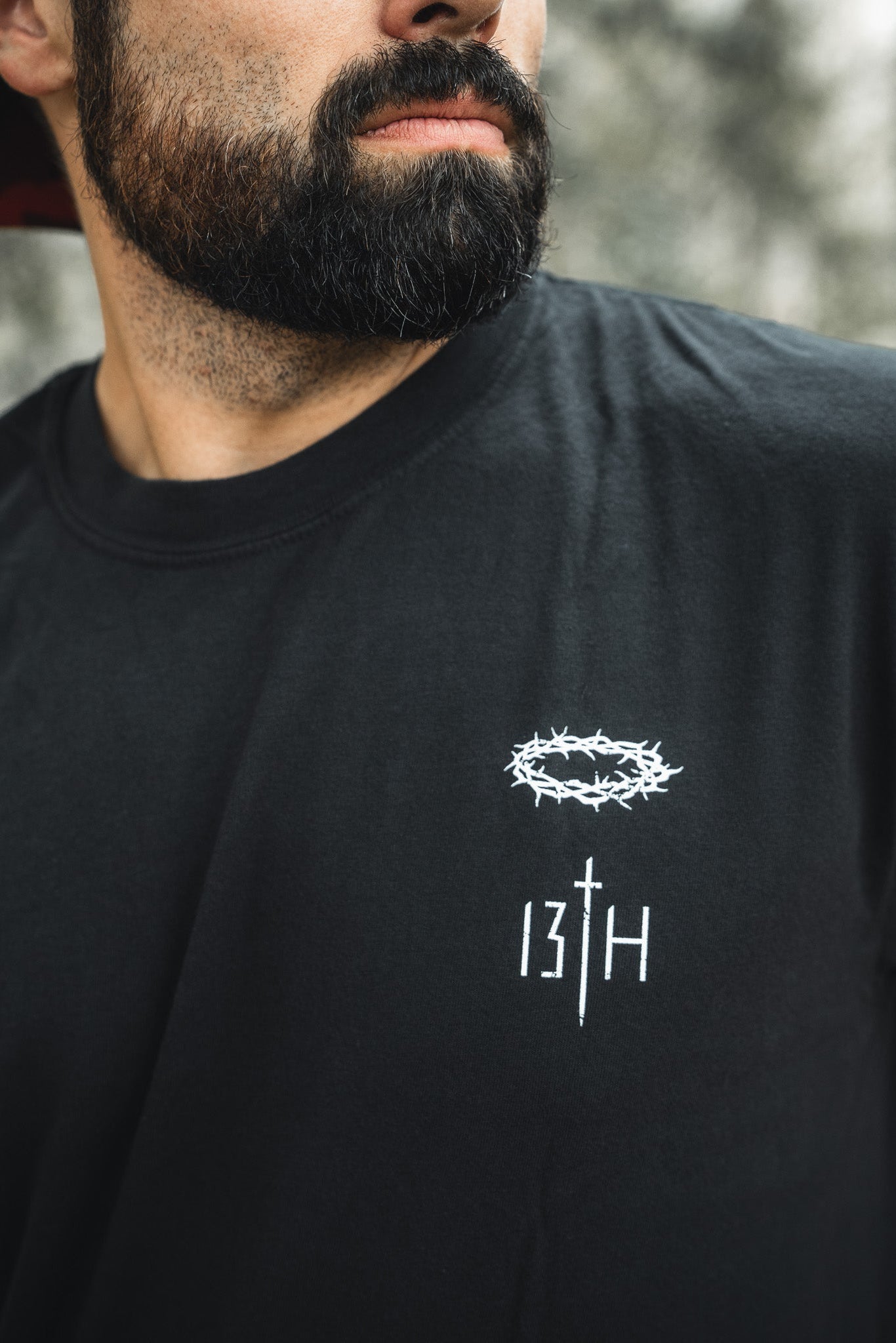 A bearded man stands out against a blurred background wearing the Jesus Saves - Black Tee from 13th, featuring a crown of thorns graphic and 13TH text in white, subtly echoing themes of everlasting life.
