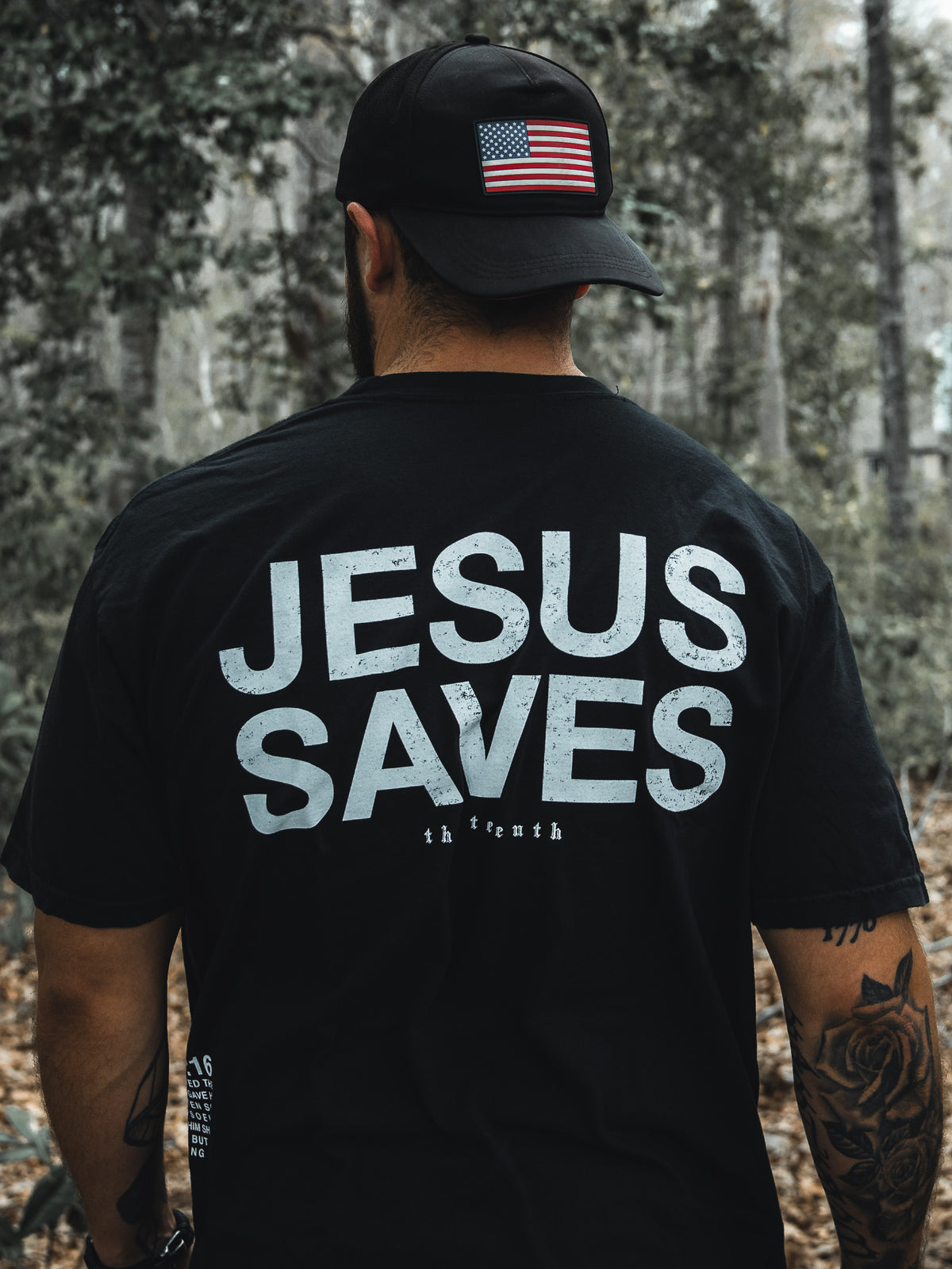In a wooded area, a man dons the Jesus Saves - Black Tee by 13th and a black cap with an American flag. His tattooed arms suggest his deep faith, possibly inspired by John 3:16s promise of everlasting life through the begotten son.