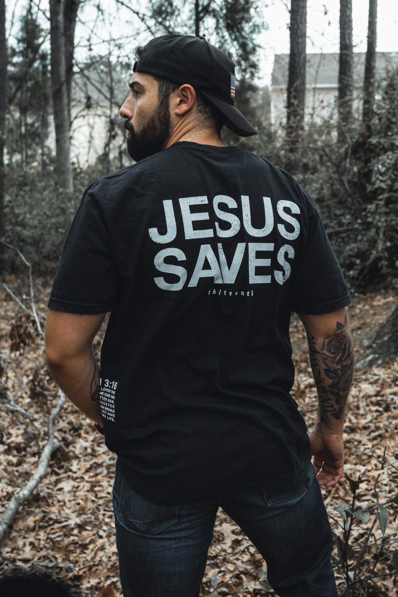 In a forest setting, a bearded man dons the 13th brands Jesus Saves - Black Tee featuring JESUS SAVES on the back. His sideways gaze amidst towering trees and leafy ground reflects the John 3:16-inspired message of eternal life.