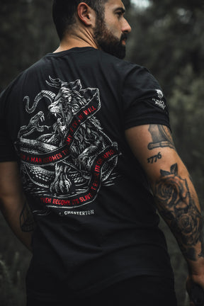 A tattooed man stands outdoors in the Born United Private Label "The Last Stand" tee, featuring a lion battling a serpent and a red-quoted banner, with a blurred green background evoking festival vibes.