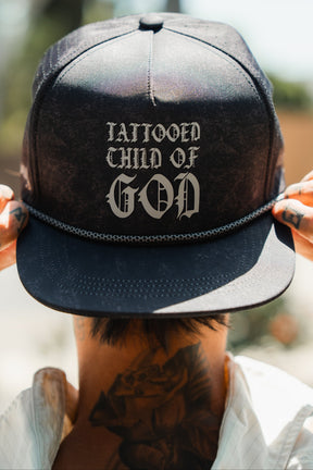 A person with neck and finger tattoos adjusts the Relentless Betrayal Tattooed Child of God Mineral Wash PVC Snapback, featuring bold silver gothic lettering, while the hat stands out against a blurred background.