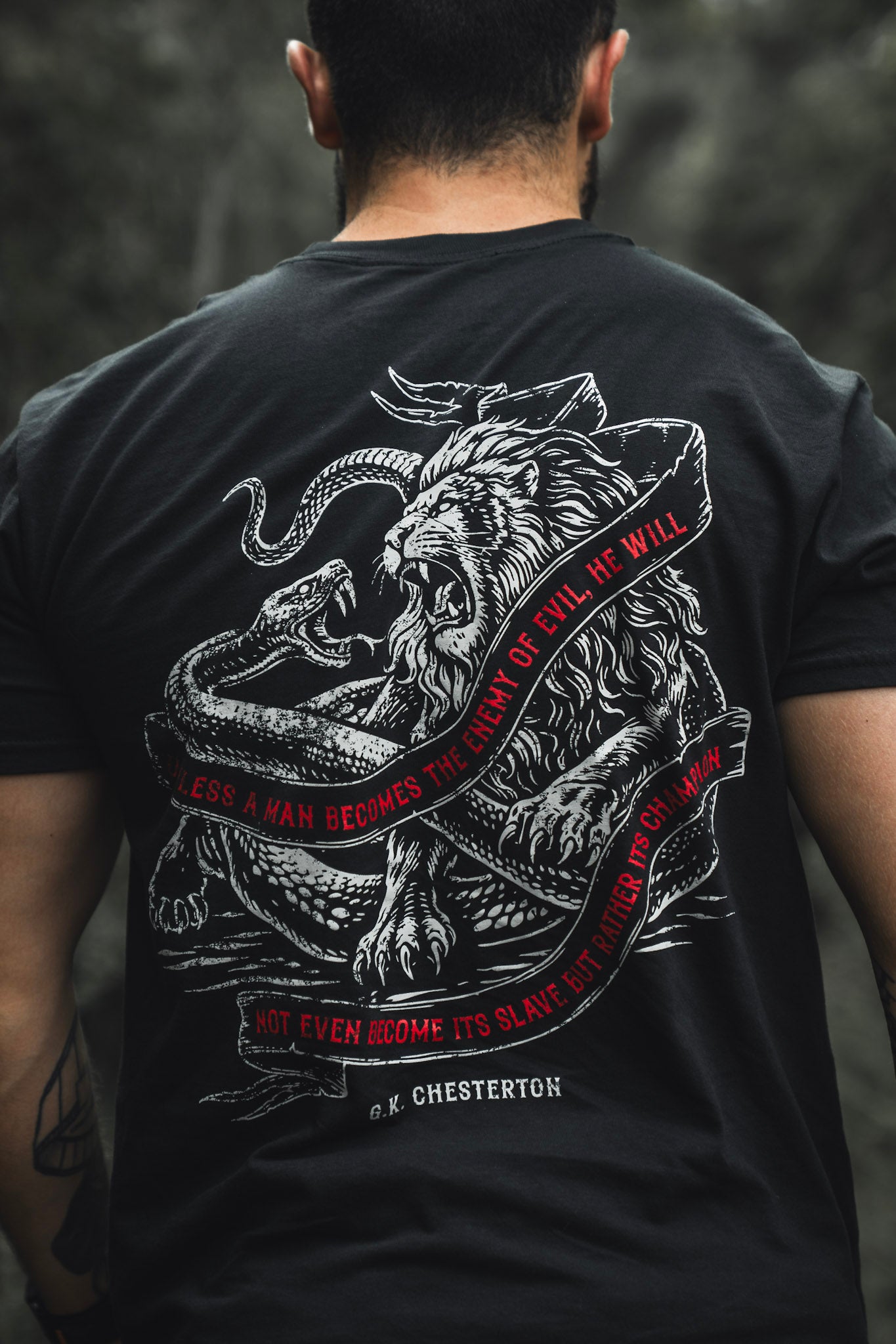 A man wears The Last Stand by Born United Private Label: a black limited edition tee with a white lion vs. serpent graphic and red text, "Unless a man becomes the enemy of evil, he will not even become its slave but rather its champion.
