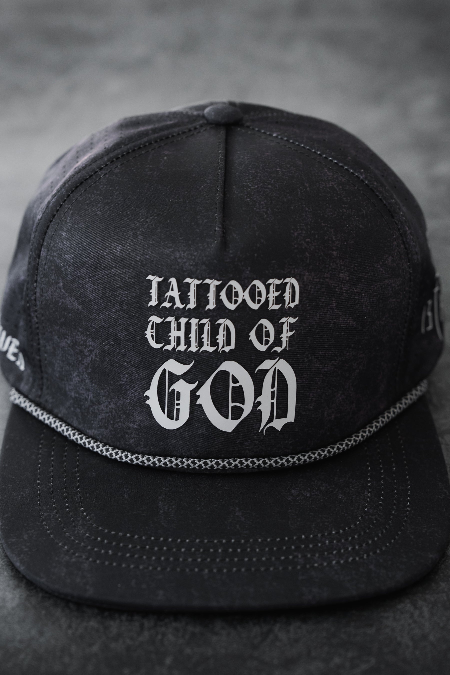 The Relentless Betrayal Tattooed Child of God Mineral Wash PVC Snapback features white gothic lettering, black mineral-washed fabric, a braided rope above the visor, and an adjustable snapback fit.