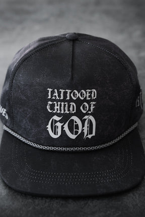 The Relentless Betrayal Tattooed Child of God Mineral Wash PVC Snapback features white gothic lettering, black mineral-washed fabric, a braided rope above the visor, and an adjustable snapback fit.