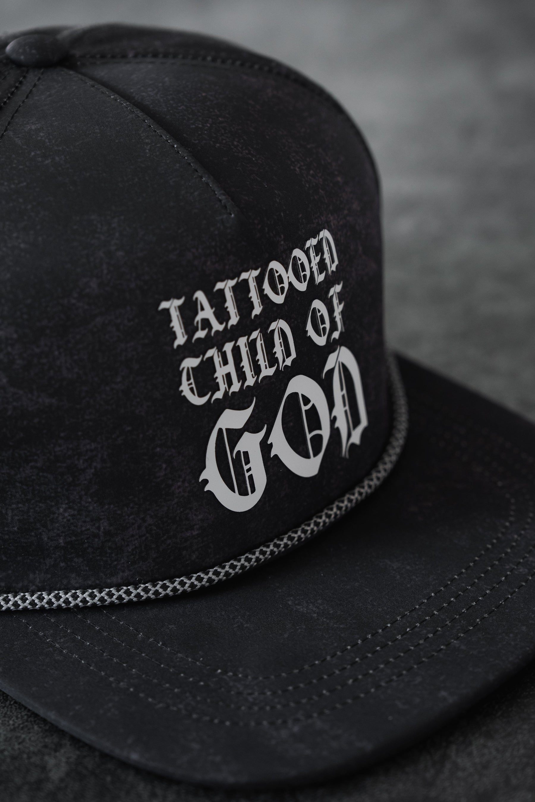 A close-up of the Relentless Betrayal Tattooed Child of God Mineral Wash PVC Snapback, showcasing white gothic "TATTOOED CHILD OF GOD" lettering and decorative stitching on the brim.