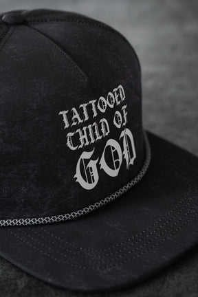 A close-up of the Relentless Betrayal Tattooed Child of God Mineral Wash PVC Snapback, showcasing white gothic "TATTOOED CHILD OF GOD" lettering and decorative stitching on the brim.