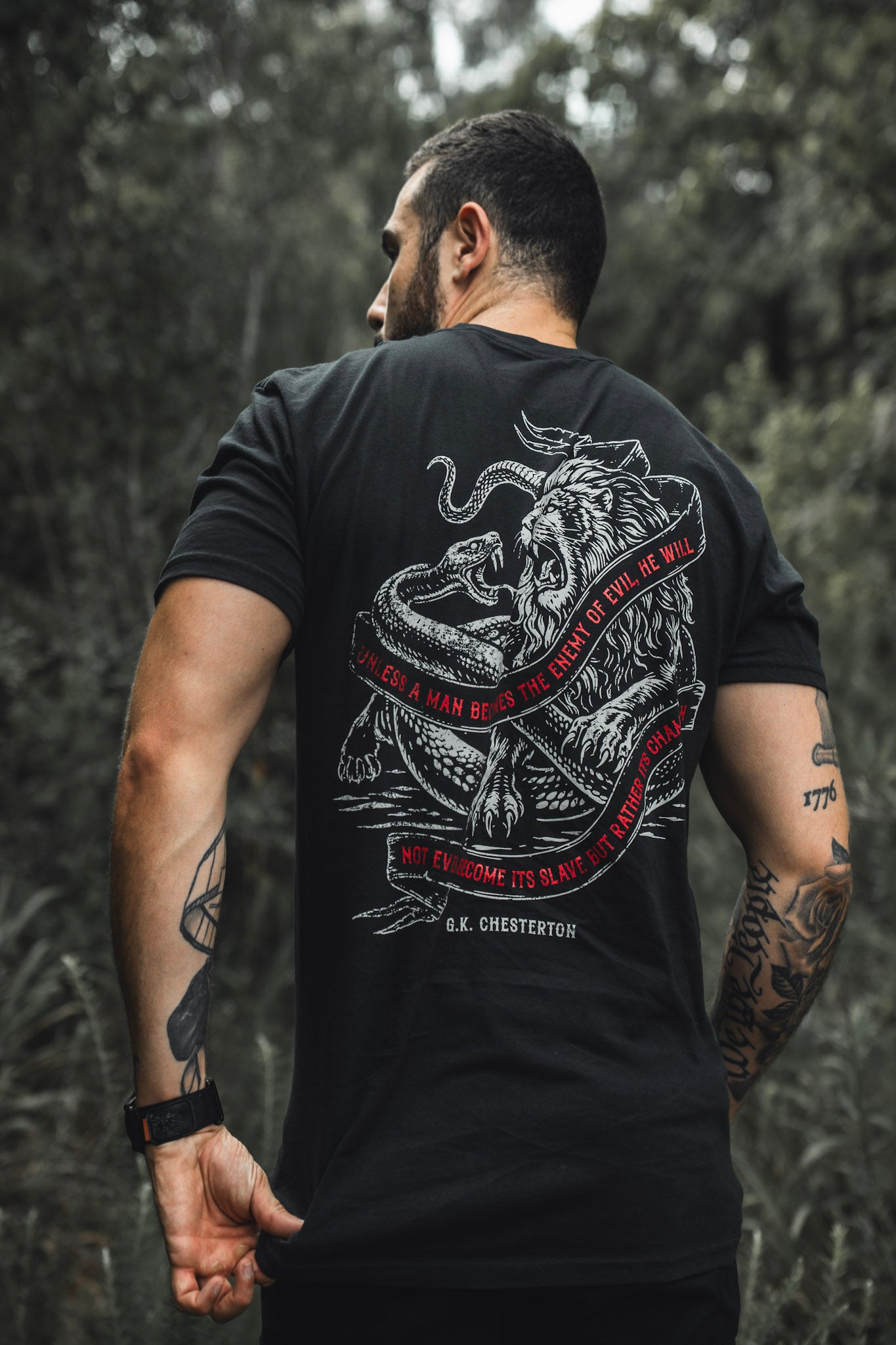 A tattooed man stands outdoors, facing away, wearing The Last Stand tee by Born United Private Label—black with a lion and snake graphic, red circular border, and G.K. Chesterton quote. Blurred trees fill the background.