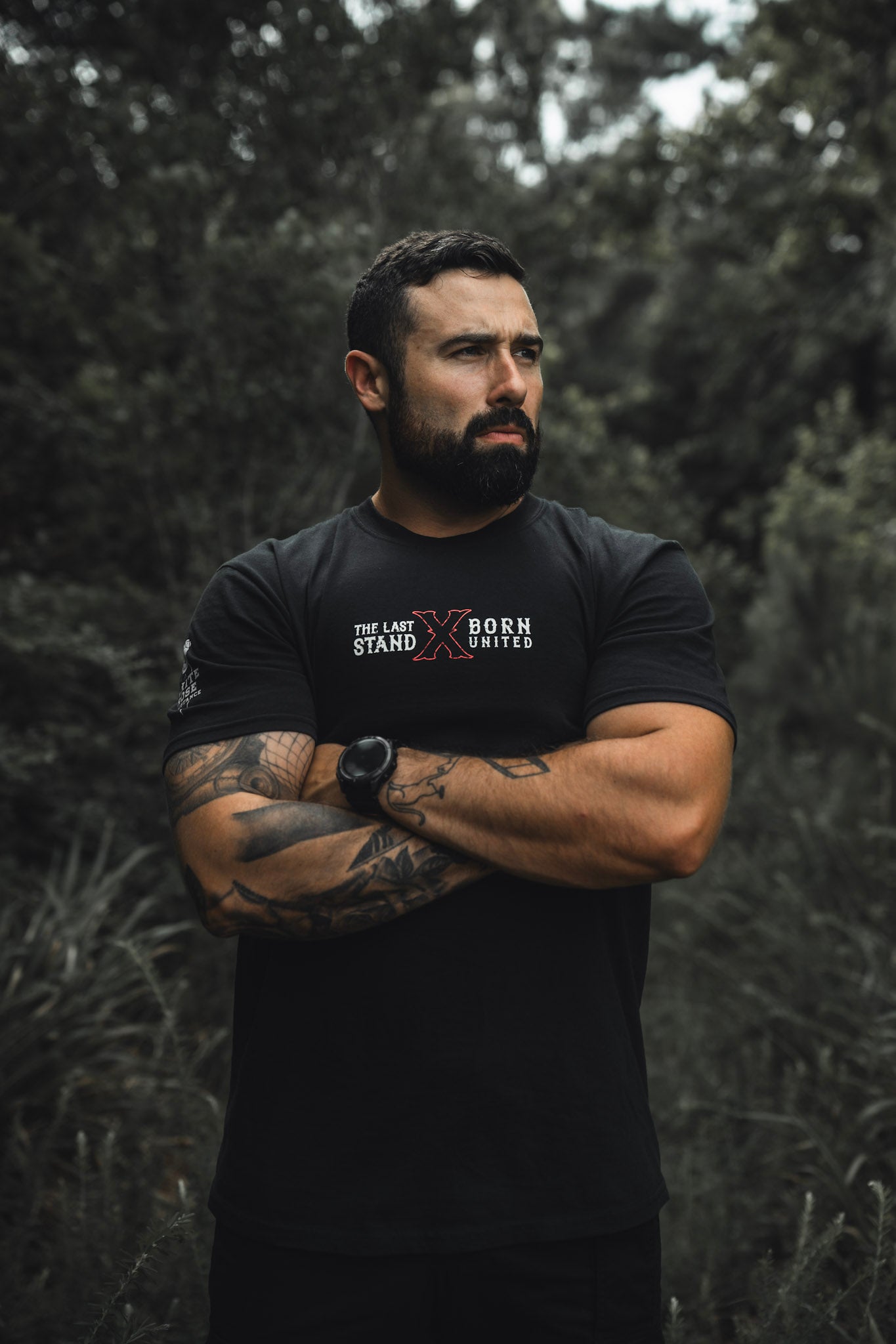 A tattooed, muscular man stands outdoors among greenery, arms crossed and looking to the side. He wears "The Last Stand" tee by Born United Private Label and a matching watch.