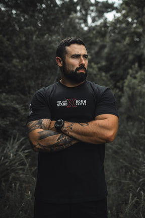 A tattooed, muscular man stands outdoors among greenery, arms crossed and looking to the side. He wears "The Last Stand" tee by Born United Private Label and a matching watch.