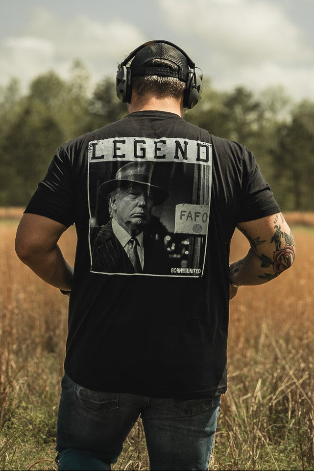 A man with headphones, sporting Born United Private Labels Legend FAFO black graphic T-shirt, strolls through a grassy field under a partly cloudy sky with trees in the background.