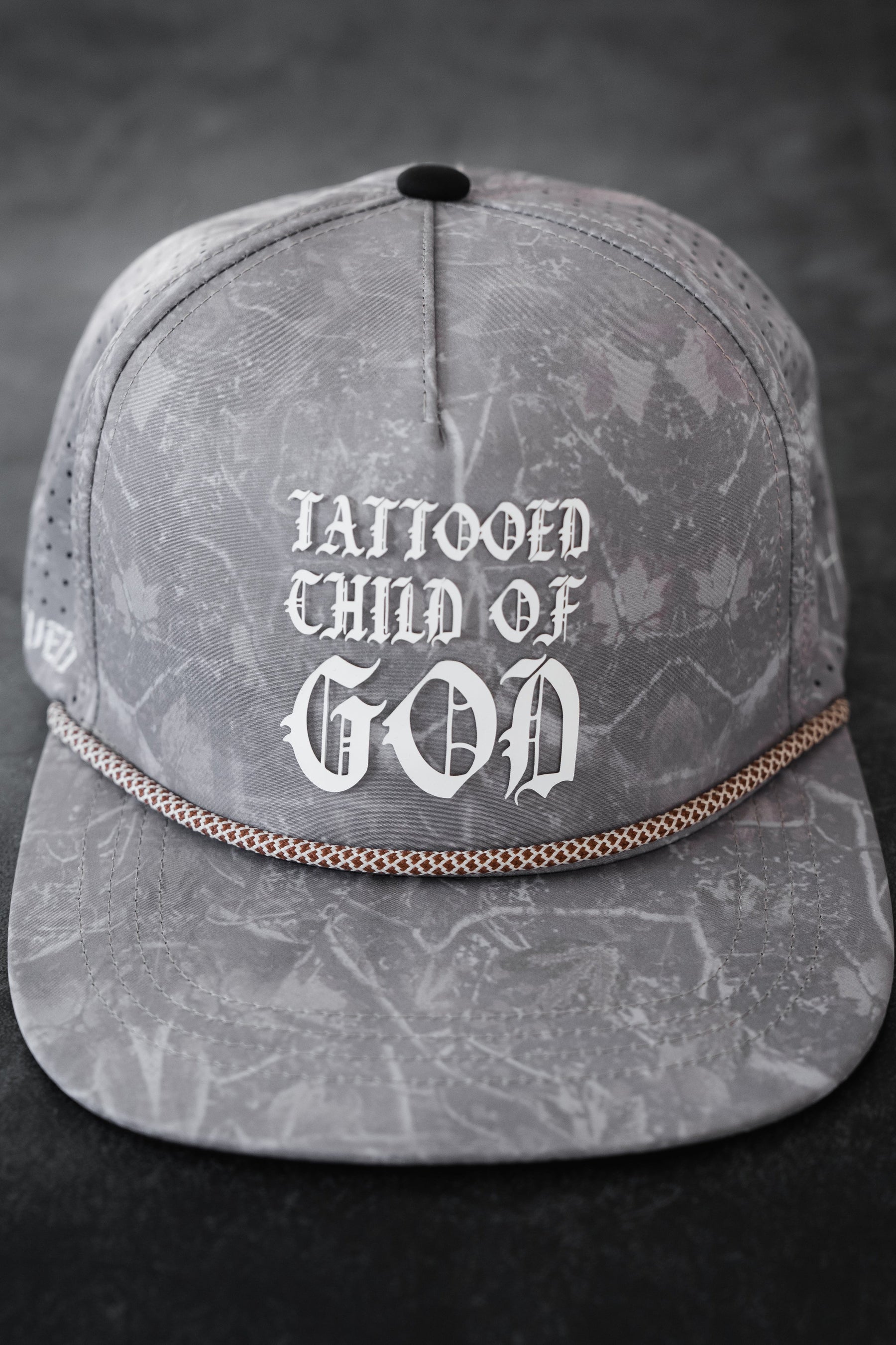 The Relentless Betrayal Tattooed Child of God Deadwood Camo PVC Snapback features a gray marbled camo design, bold white Gothic-style "TATTOOED CHILD OF GOD" text on the front, and a decorative chain on the brim.