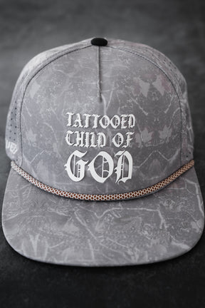 The Relentless Betrayal Tattooed Child of God Deadwood Camo PVC Snapback features a gray marbled camo design, bold white Gothic-style "TATTOOED CHILD OF GOD" text on the front, and a decorative chain on the brim.