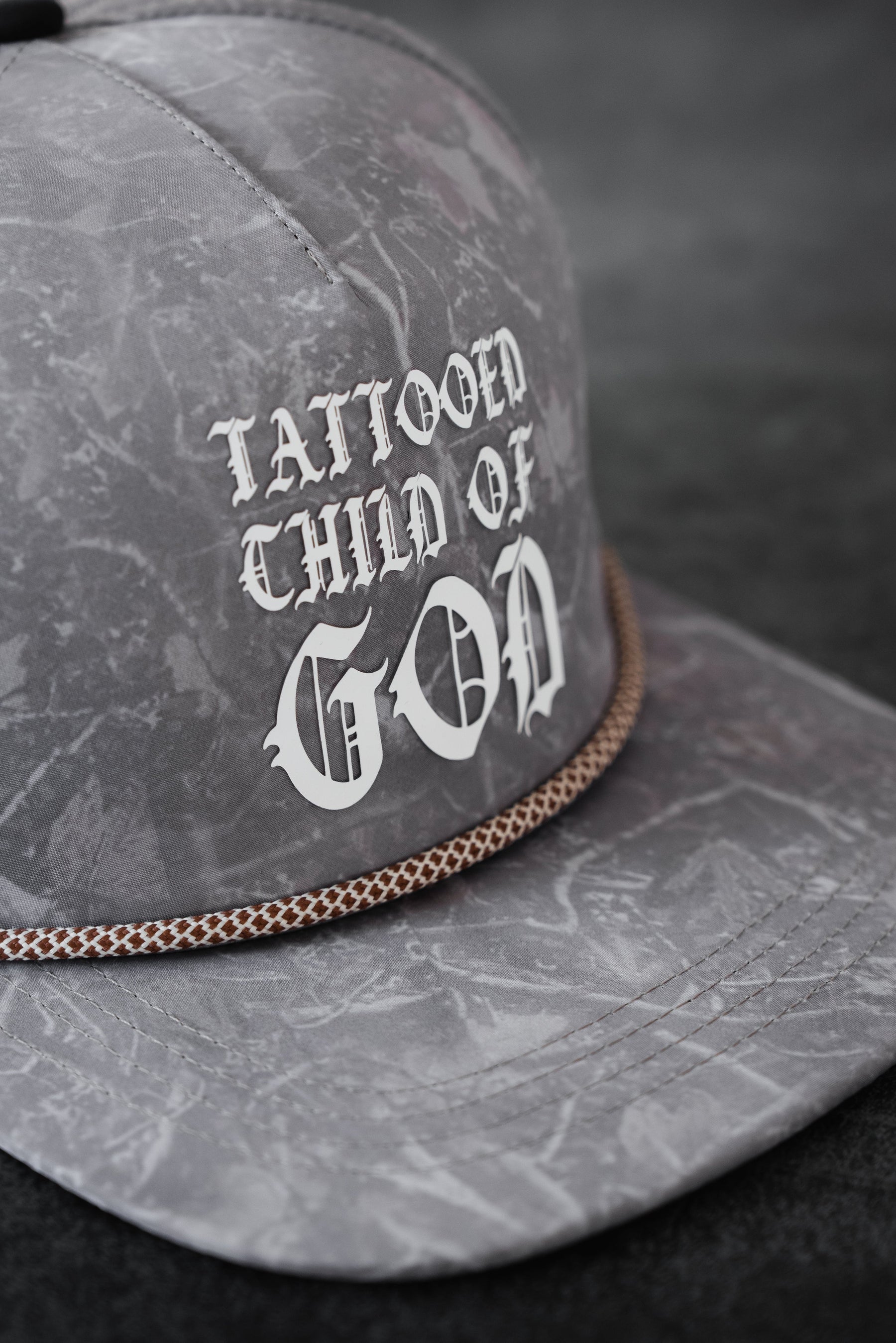 The Relentless Betrayal Tattooed Child of God Deadwood Camo PVC Snapback features white gothic lettering and a braided brim, making it bold Christian apparel for any collection.