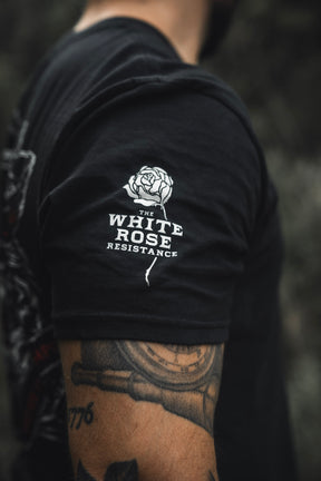 A person wears The Last Stand tee by Born United Private Label, a black shirt with a white rose graphic and THE WHITE ROSE RESISTANCE on the sleeve. Their arm tattoo stands out against the blurred outdoor background at The Last Stand Festival.