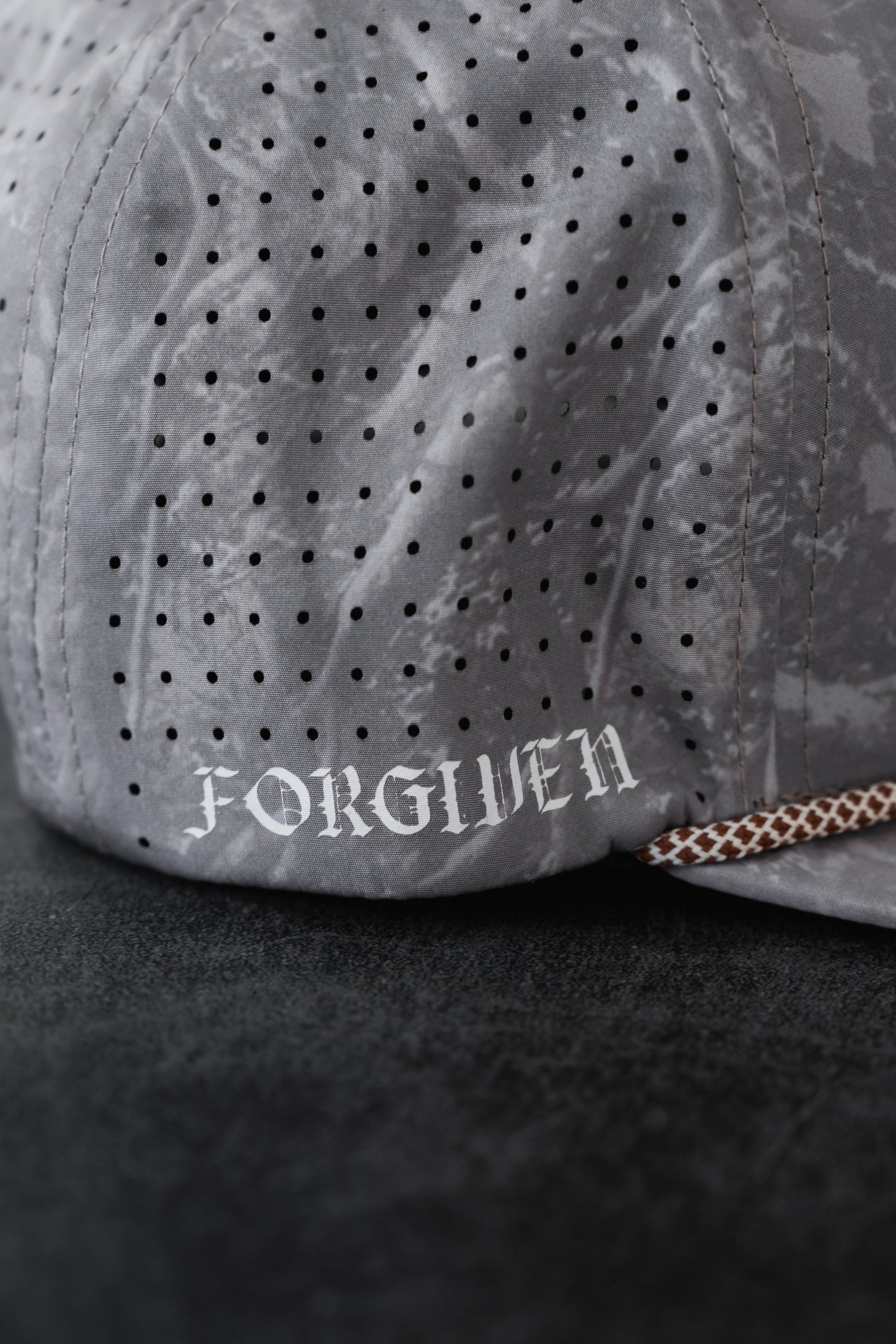 A close-up of the Tattooed Child of God Deadwood Camo PVC Snapback by Relentless Betrayal, a gray perforated cap with white gothic FORGIVEN text on the side, inspired by Christian apparel and resting on a dark surface.