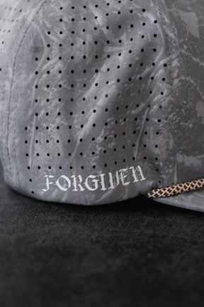 A close-up of the Tattooed Child of God Deadwood Camo PVC Snapback by Relentless Betrayal, a gray perforated cap with white gothic FORGIVEN text on the side, inspired by Christian apparel and resting on a dark surface.