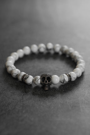 The Until Death Storm Cloud Bead Bracelet by Relentless Betrayal showcases white marbled beads and a bold metallic skull centerpiece, stylishly presented on a dark, textured surface.
